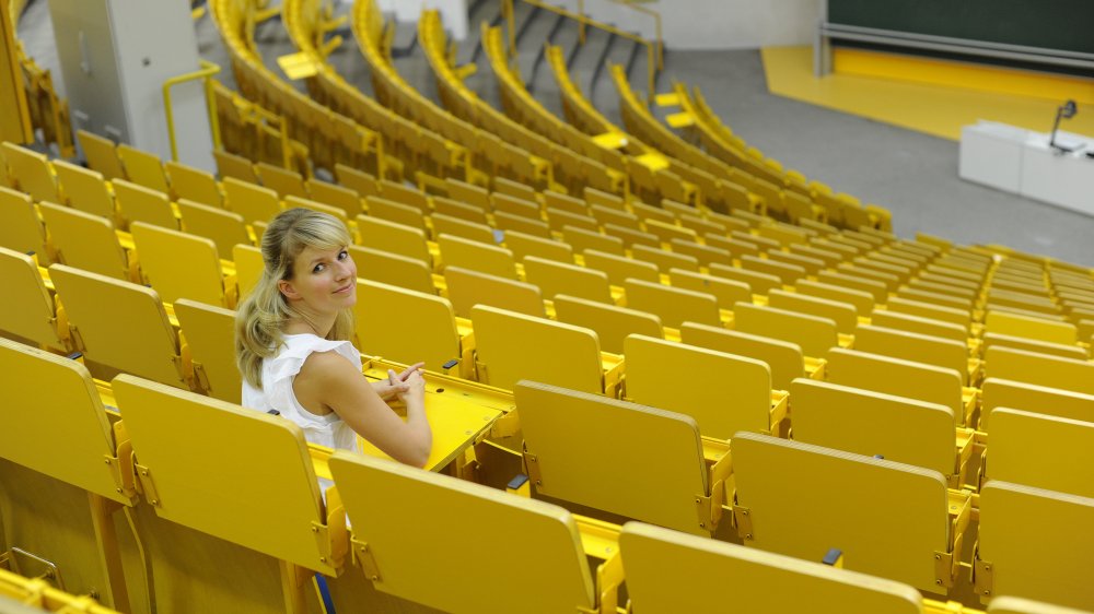 A young woman is sitting in an lecture hall with yellow chairs. 