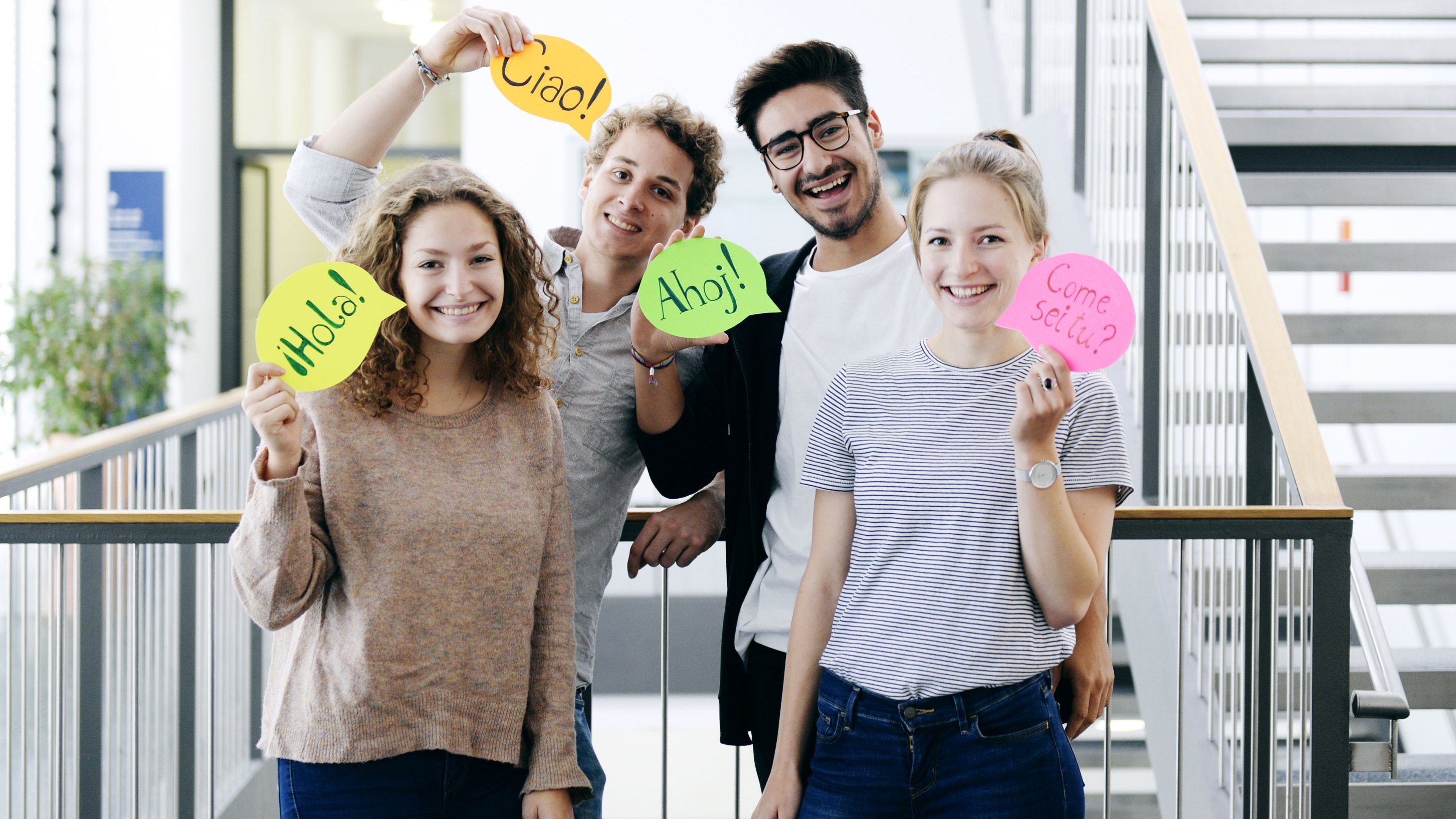 A group of four young people show cards that are formed like speech bubbles. Each of them says hello in a different language.