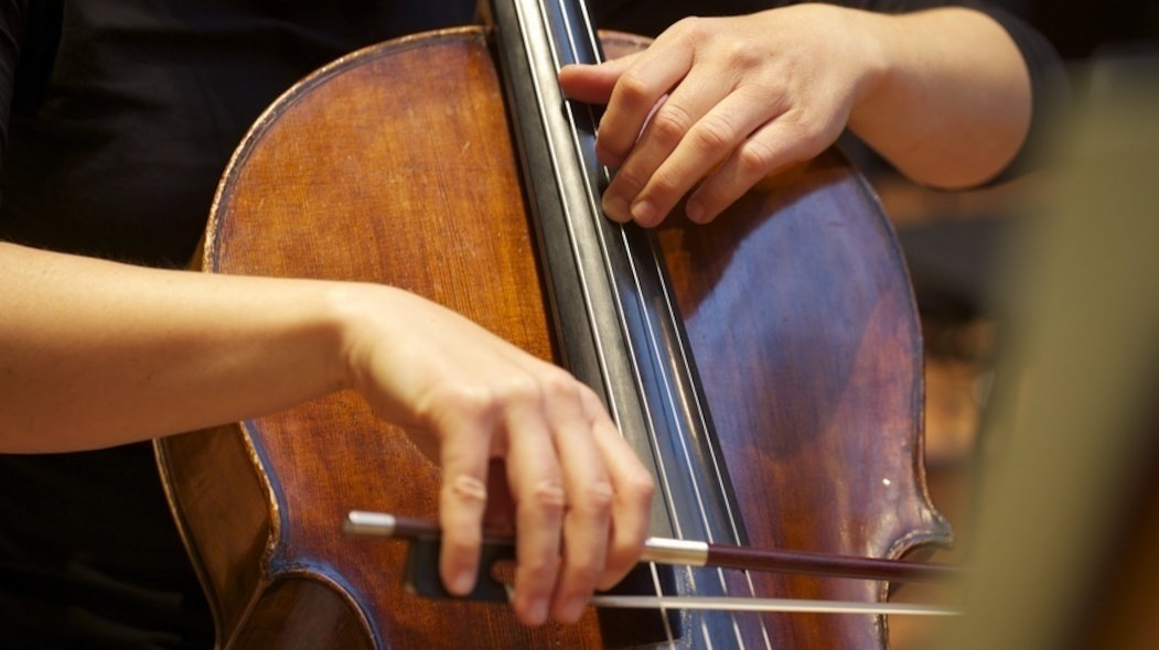 Nahaufnahme eines Cellos, das von einem Mann gehalten wird. Eine Hand hält einen Bogen über das Instrument. Die andere hält die Saiten fest.