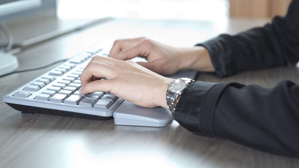 Zwei Hände einer Person schreiben auf einer silbernen Tastatur, die auf einem Tisch vor einem Monitor steht.