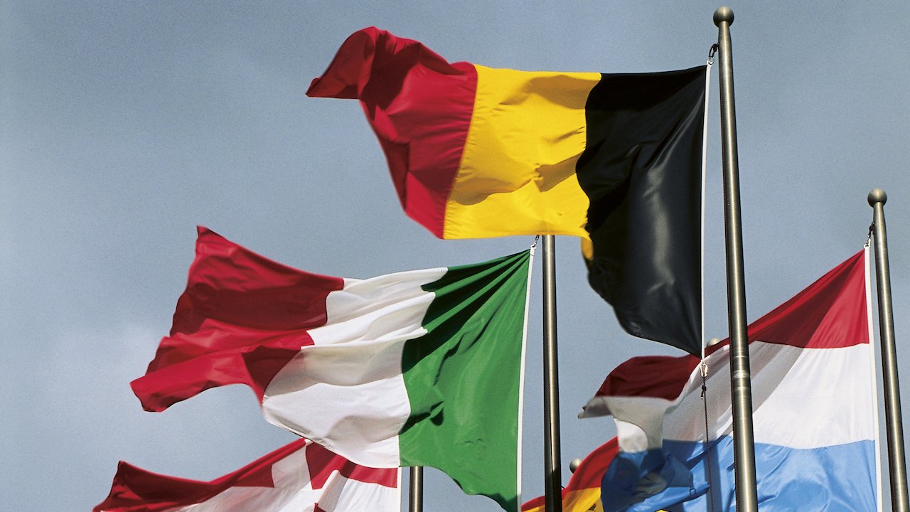 Aufgenommen aus der Froschperspektive ragen neben einer Europafahne verschiedene Flaggen von europäischen Mitgliedsstaaten in den bewölkten Himmel.