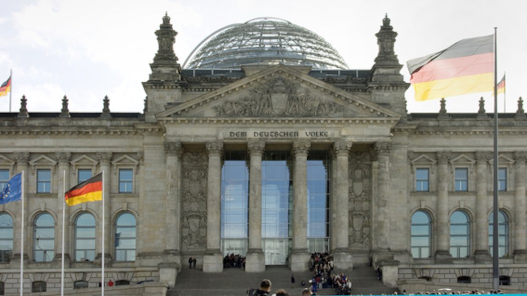 Aufnahme des Reichstagsgebäudes mit Glaskuppel in Berlin. Auf und vor dem Gebäude stehen mehrere Deuschlandflaggen und eine Europaflagge.