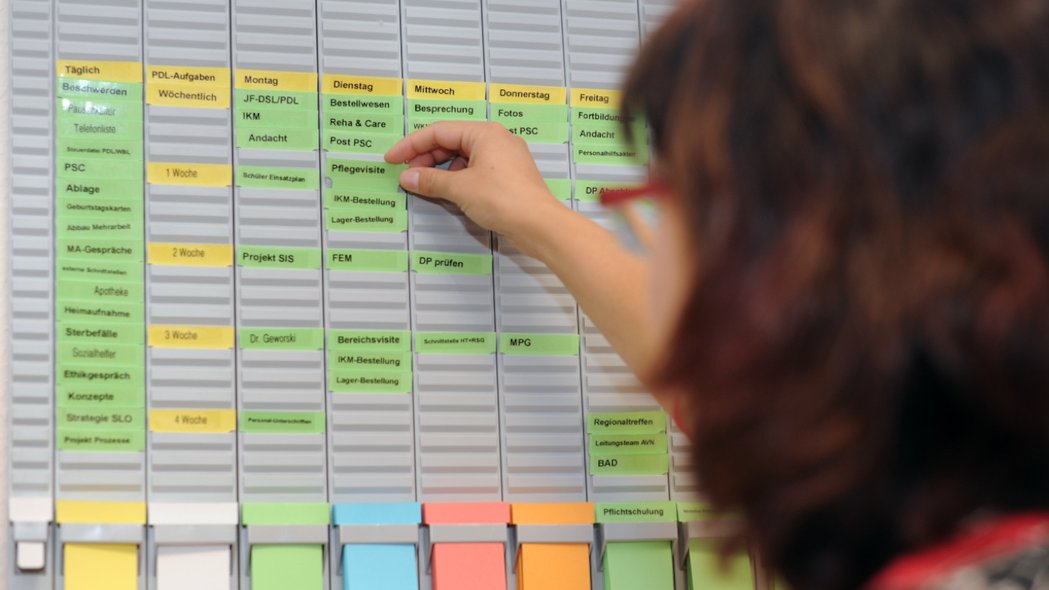 Eine Frau hält ein Schild mit der Aufschrift Pflegevisite in einen Wochenplan, der an einer Wand hängt.