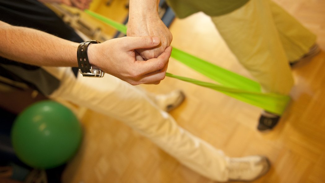 Auf hellem Holzboden sieht man zwei männliche Personen brustabwärts. Die eine Person wird von der anderen bei einer Übung mit einem leuchtend grünen Fitnessband angeleitet.