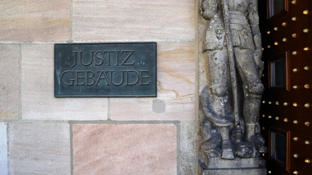 An der rosafarbenen Sandsteinmauer neben dem Gebäudeeingang ist eine dunkle Metallplakette mit der Aufschrift "Justiz Gebäude" angebracht. Daneben eine graue Steinstatue.