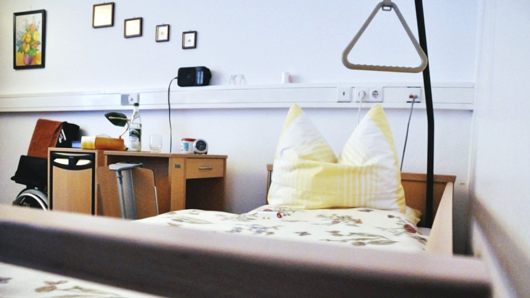 Ein Krankenbett aufgenommen vom unteren Ende. Neben dem Krankenbett steht ein Nachtschrank mit ausklappbarem Tablett. An der Wand hängen Bilder in verschiedenen Größen.