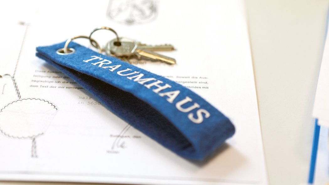 Auf einem notariellen Schriftstück liegt ein Schlüssel mit einem blauen Schlüsselanhänger aus Filz mit der Aufschrift Traumhaus.