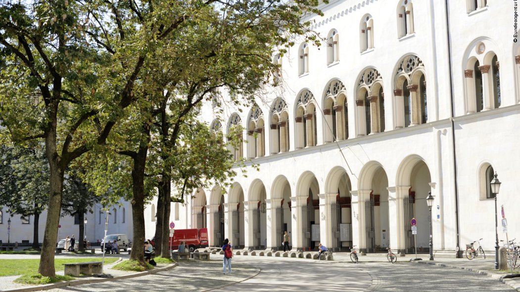 Blick auf ein weißes Gebäude in der Sonne mit Säulen und Bogengängen gegenüber von einem Platz mit Bäumen und Sitzgelegenheiten.