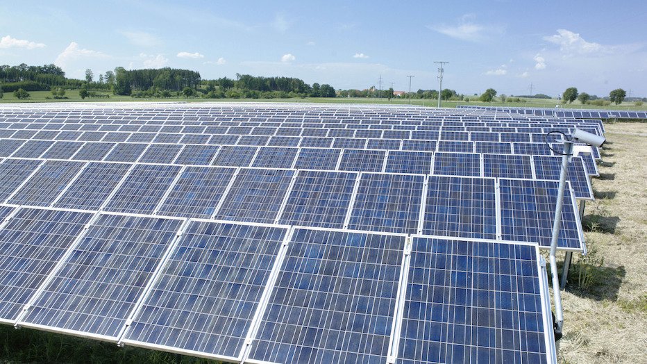 Blick über ein Feld auf dem eine Solaranlage installiert wurde. Im Hintergrund sind Büsche, Bäume und Felder zu sehen.