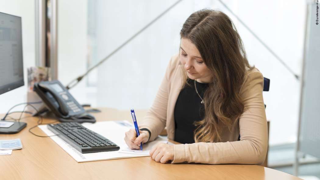 Eine Bankkauffrau sitzt am Schreibtisch und schreibt mit einem Stift.