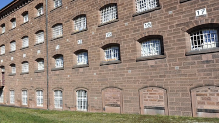 Hinter einem Grünstreifen steht ein gemauertes Gebäude mit weißen Gittern vor den Fenstern. Am Gebäude sind Zahlen aufgemalt.
