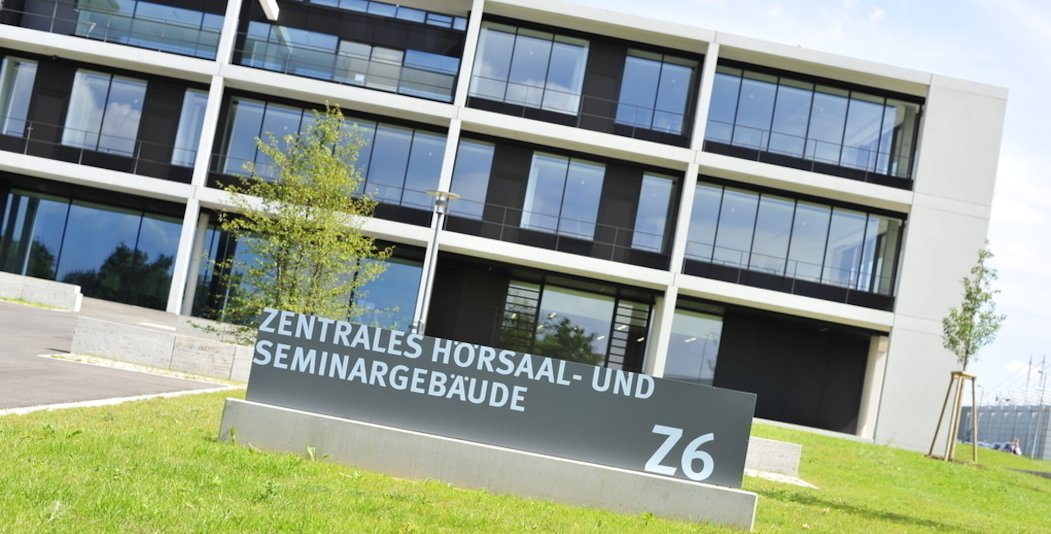 Außenaufnahme eines modernen dreistöckigen Gebäudes auf einer grünen Wiese. Vor dem Gebäude steht ein Schild mit der Aufschrift: Zentrales Hörsaal- und Seminargebäude.