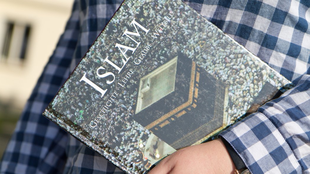 Nahaufnahme eines Buches über den Islam, welches sich eine Person unter den linken Arm geklemmt hat.