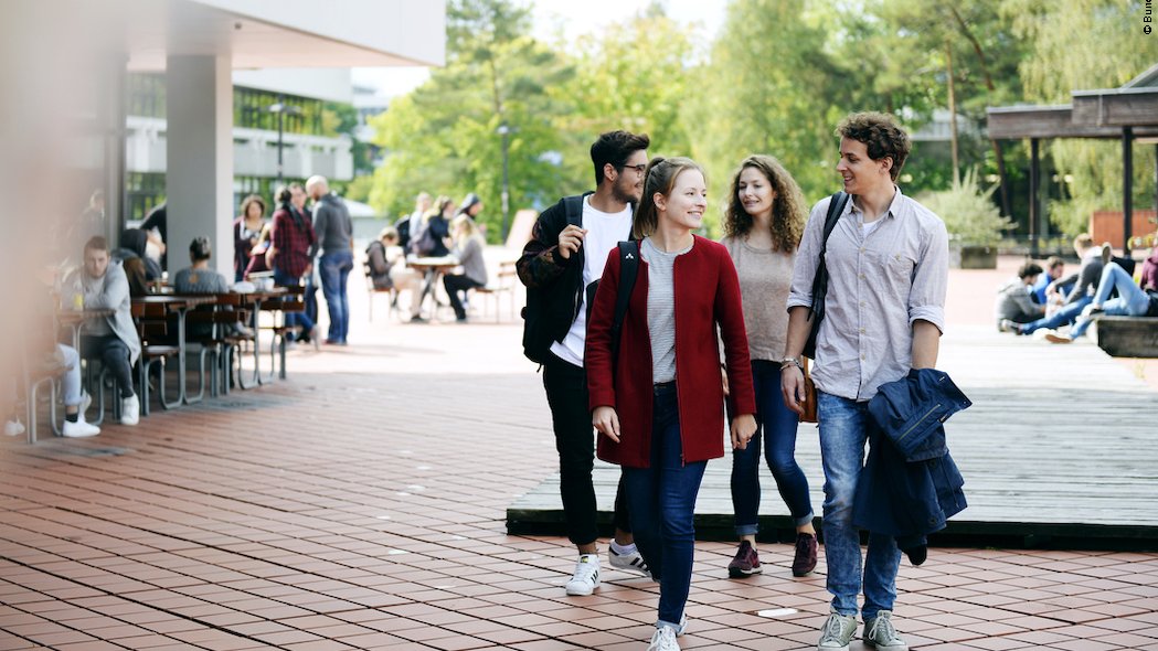 Zwei Studentinnen und zwei Studenten gehen durch einen Hochschulcampus und unterhalten sich.