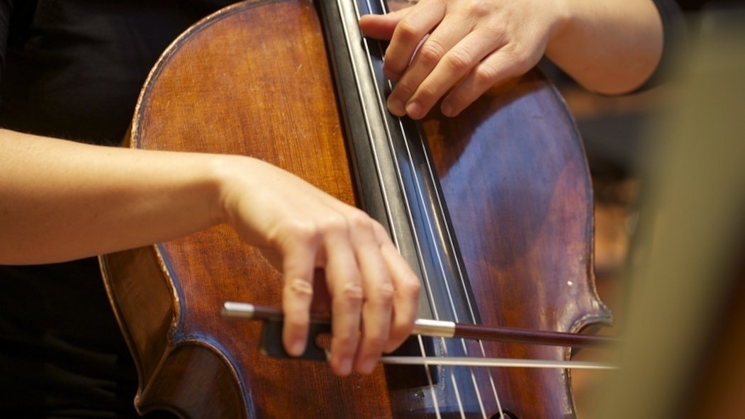 Nahaufnahme eines Cellos, das von einem Mann gehalten wird. Eine Hand hält einen Bogen über das Instrument. Die andere hält die Saiten fest.
