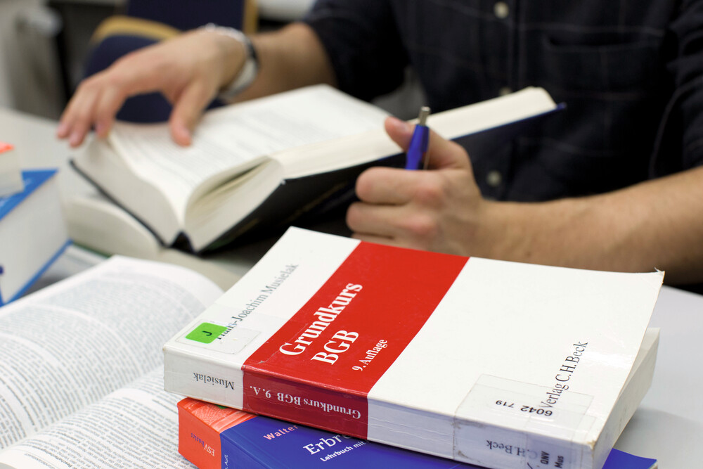 Auf einem Schreibtisch liegen mehrere verschiedene geöffnete und geschlossene Bücher neben- und auch aufeinander. Im Vordergrund liegen zwei geschlossene Bücher, deren Buchrücken zum Betrachter zeigen. Auf den obenliegenden Buchdeckel steht "Grundkurs BGB". Der Titel des darunerliegenden Buches ist nicht erkennbar. Im Hintergrund hält eine Person, die einen Stift in einer Hand hat, ein Buch aufgeschlagen. 