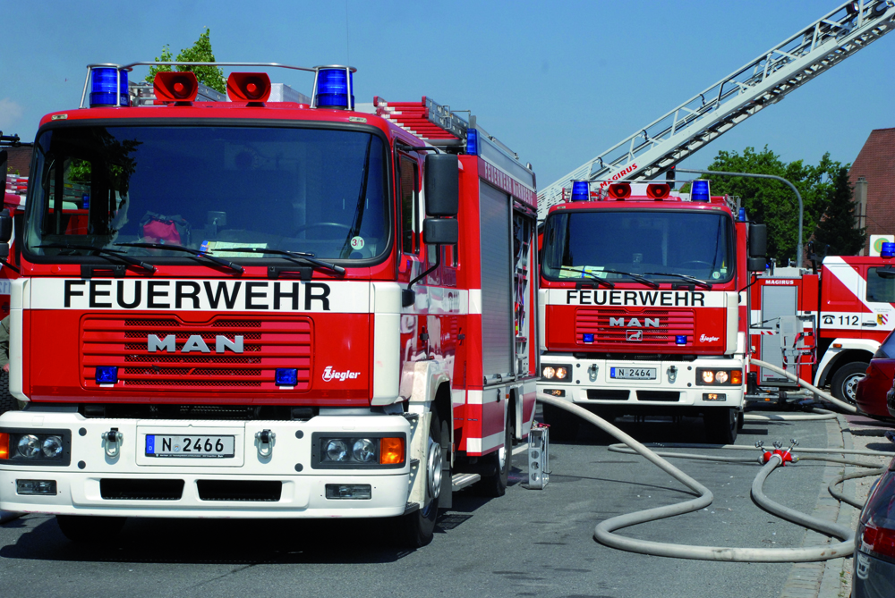Nahaufnahme von zwei Löschfahrzeugen der Feuerwehr, eines davon mit ausgefahrener Drehleiter, bei einem Einsatz in einer Wohnsiedlung. Zwei weitere Fahrzeuge im Hintergrund. Die ausgerollten Schläuche sind durch parkende Autos gelegt.